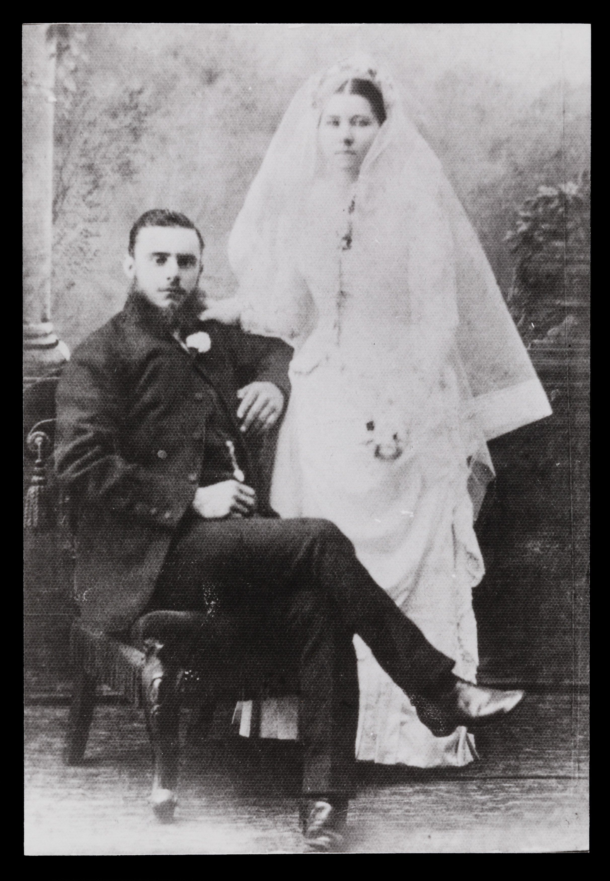 Black and white wedding photograph. The groom is wearing a suit and sitting in a chair, and beside him stands the bride in a white wedding dress and veil.